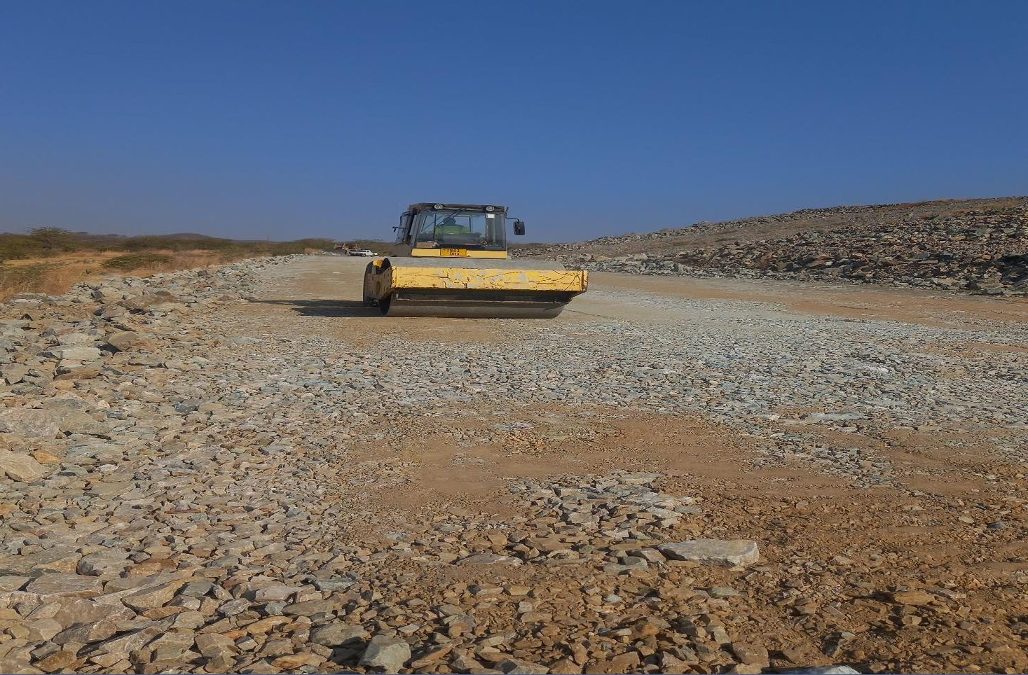 A yellow vibrating roller compacting the crest of a rockfill tailings embankment under a deep blue sky at a gold mine site