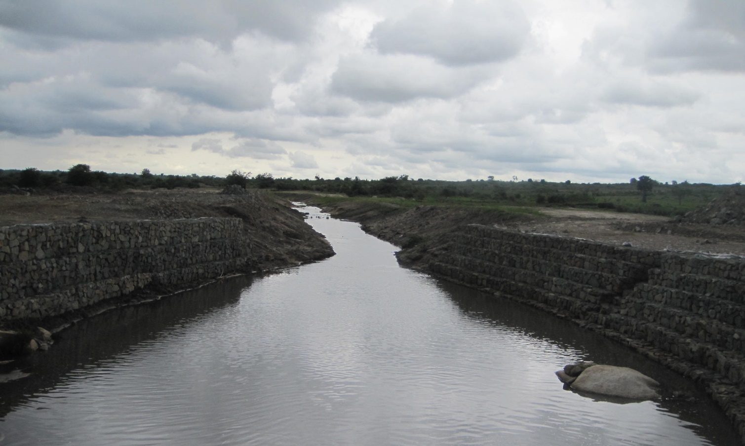 A trained river channel carrying water between two banks faced with stone-filled gabion revetment, under an overcast sky in open bush country