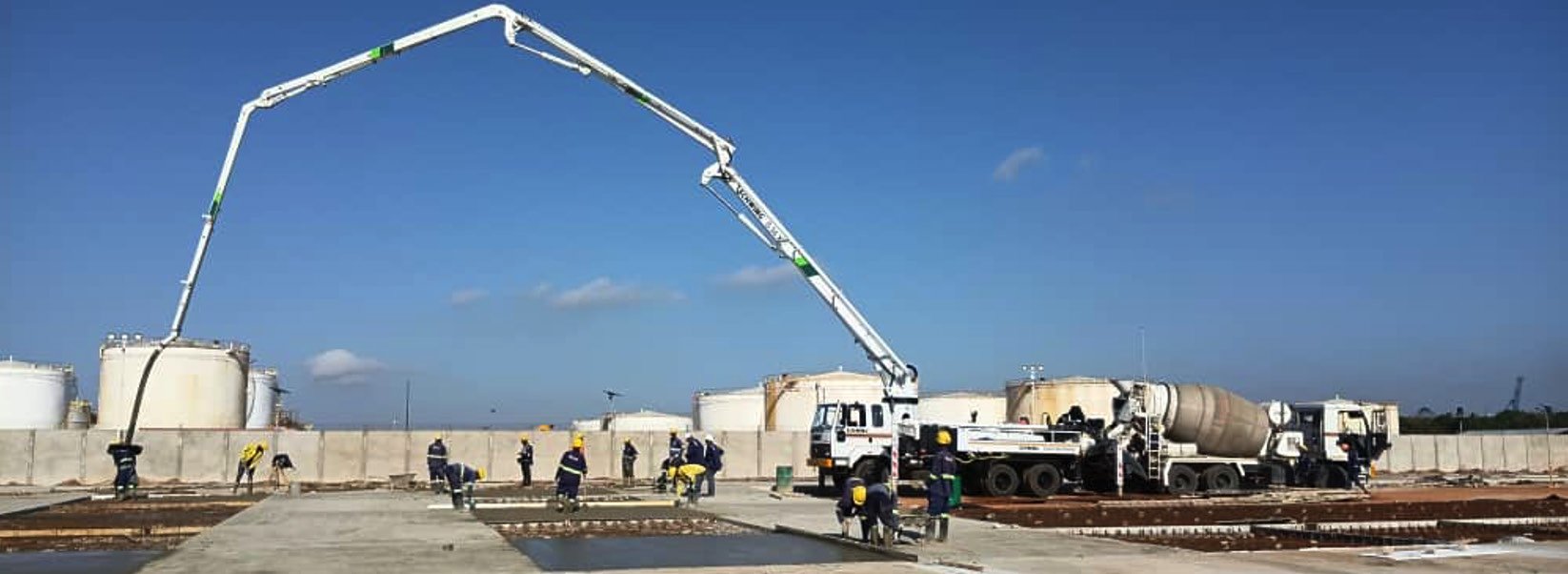 A concrete boom pump placing concrete for a rigid pavement slab inside a fuel depot, with a mixer truck, a team of workers in high-visibility vests and rows of bulk storage tanks behind