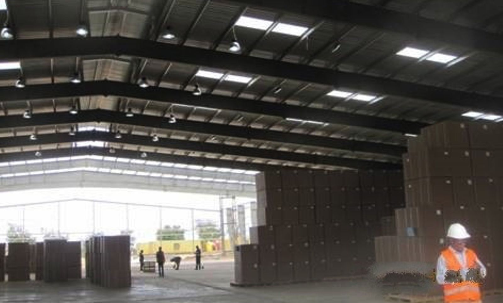 The interior of a long-span steel-framed warehouse with roof lights, stacked cartons and a worker in a hard hat and high-visibility vest in the foreground