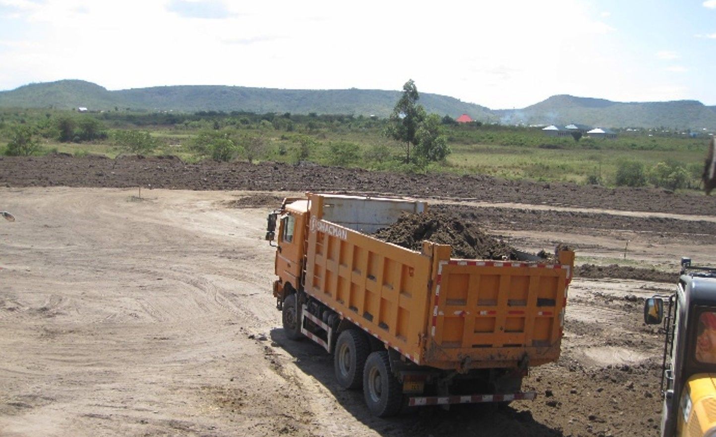 A loaded orange dump truck crossing a prepared earth platform, with hills and rooftops beyond