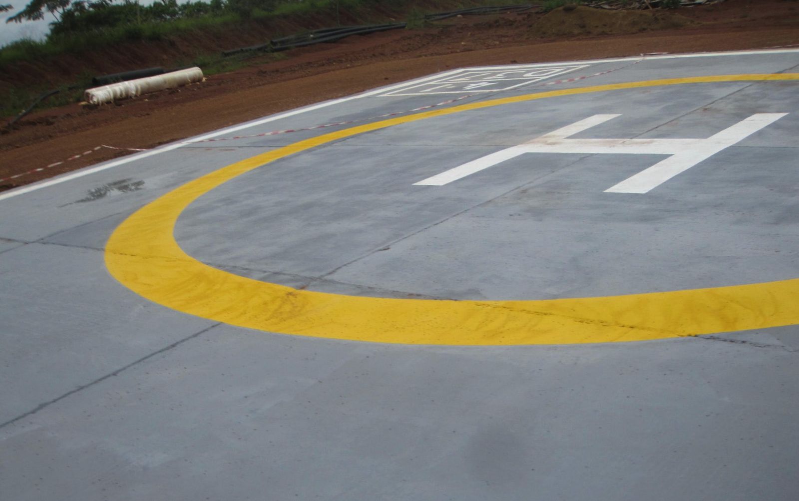 A concrete helicopter landing pad marked with a white H inside a yellow circle, with a gravel road behind