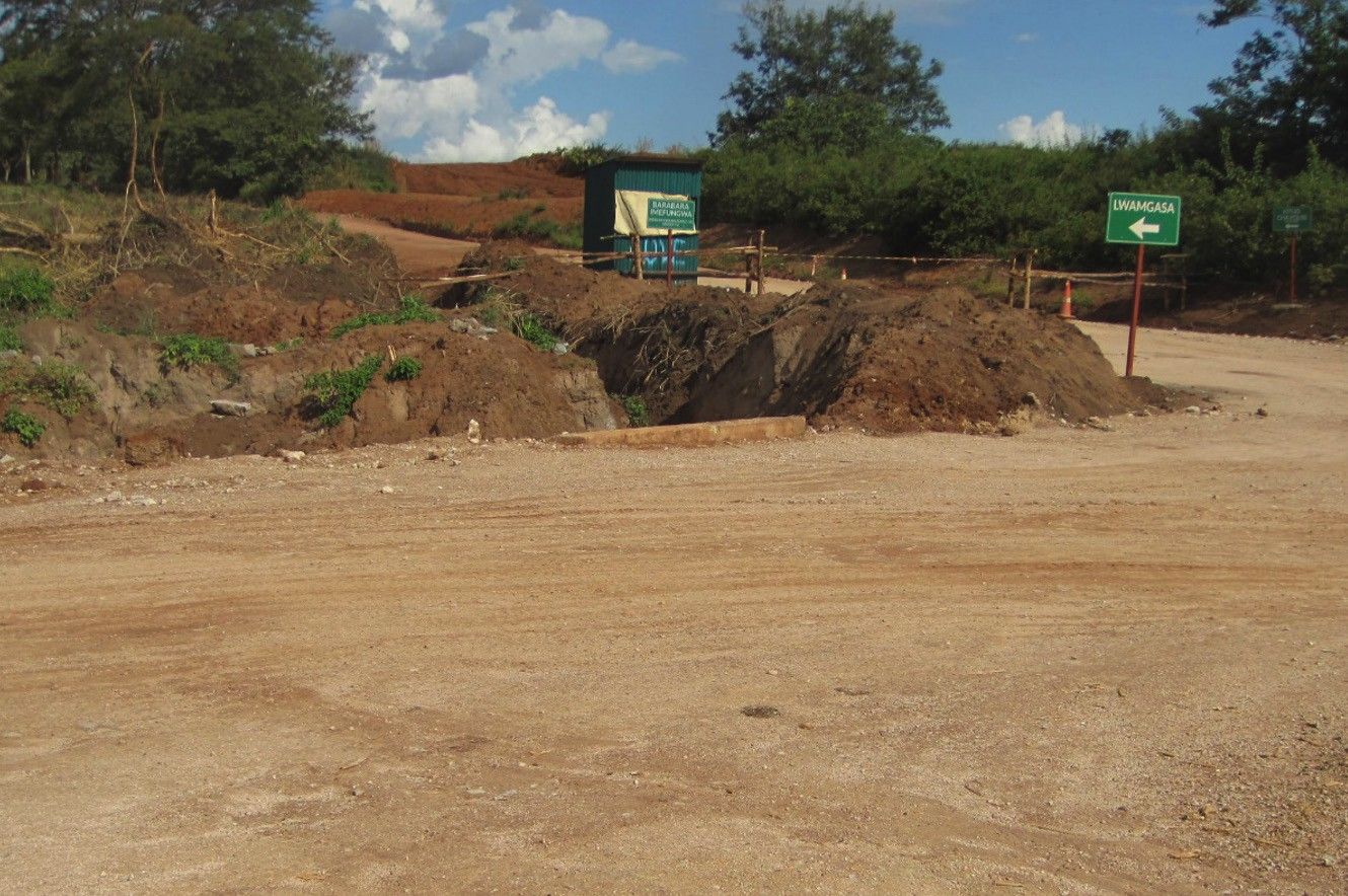 A freshly graded gravel road junction with earthworks and excavated soil, a small site office and a green direction sign at the roadside