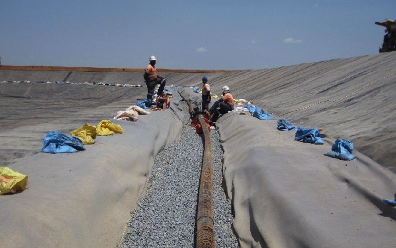 Workers laying a drainage pipe in a gravel-filled trench between geotextile-covered slopes inside a lined cell