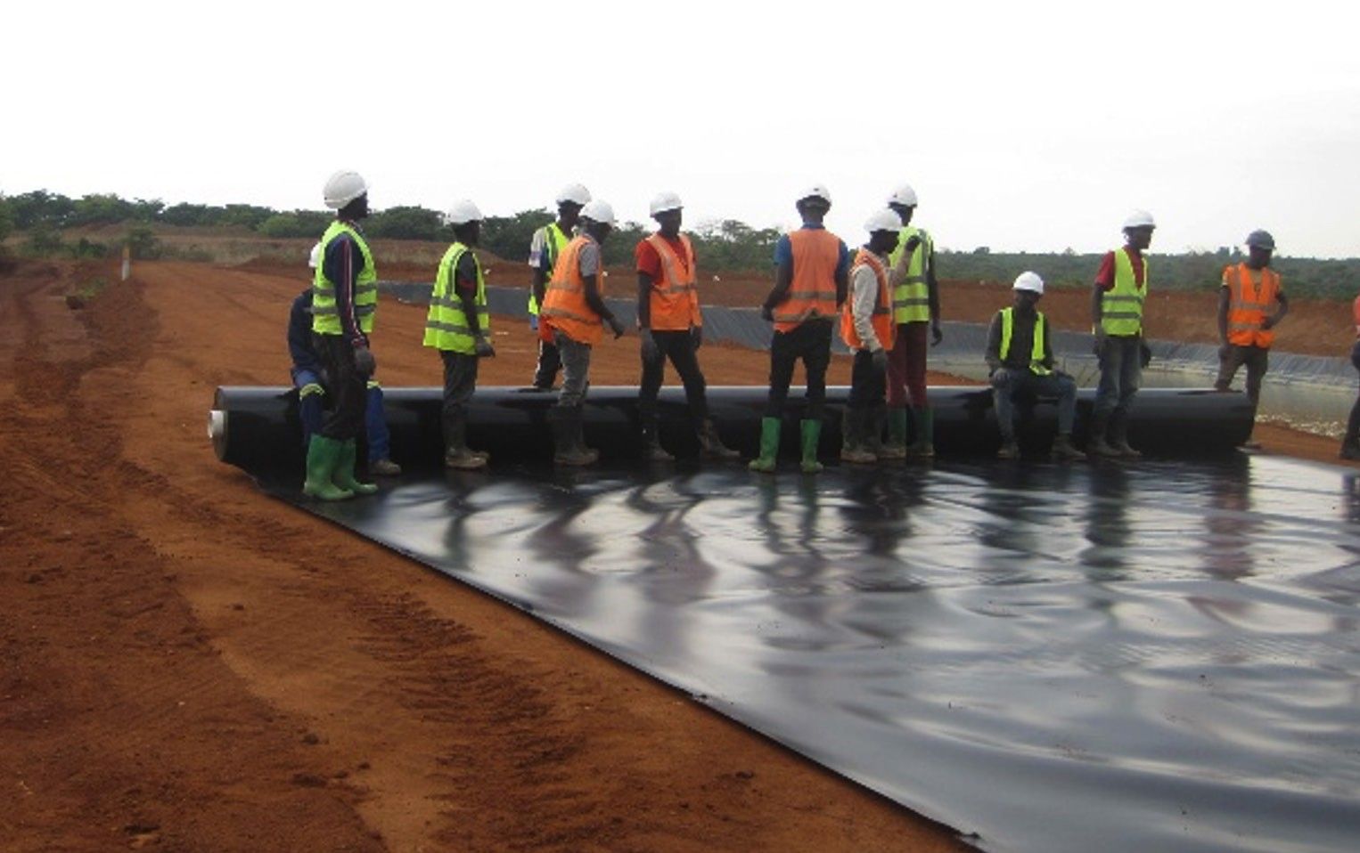 A crew of about a dozen workers in hard hats and high-visibility vests unrolling a large roll of black geomembrane liner across a prepared earthworks platform