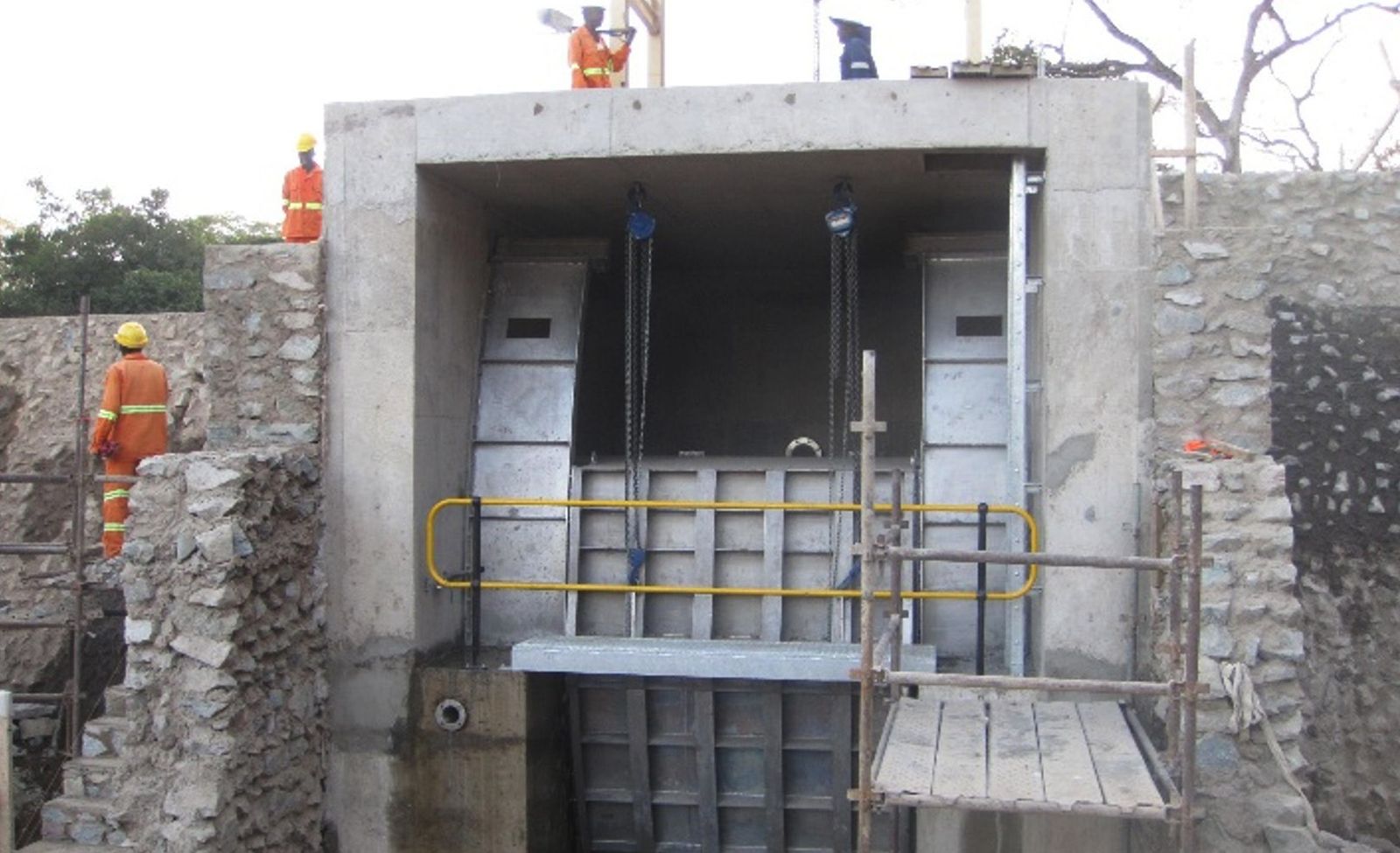 A reinforced concrete outlet structure housing two steel sluice gates with chain hoists, set into a masonry wall, with workers in orange overalls and hard hats