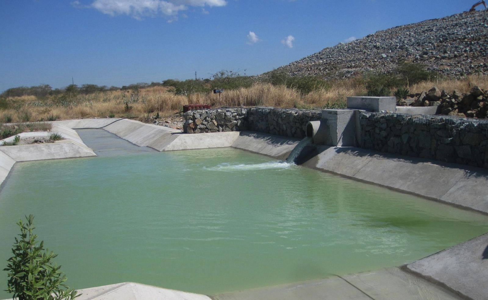 A green water-filled concrete stilling basin with gabion retaining walls and a pipe outfall, below a rockfill embankment under a blue sky