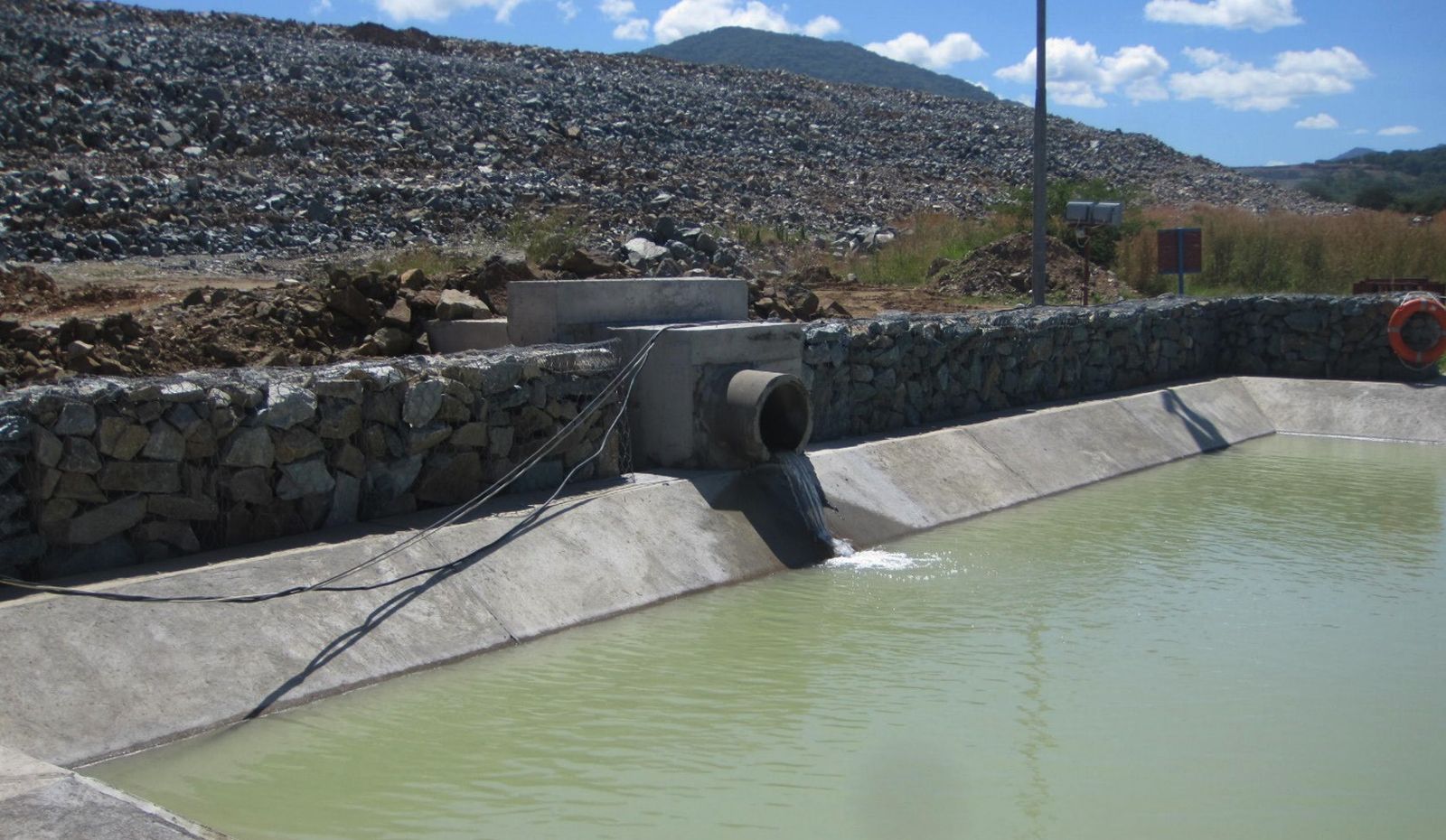 A concrete-lined stilling basin filled with water, fed by a pipe outfall discharging through a gabion retaining wall, with a rockfill embankment behind