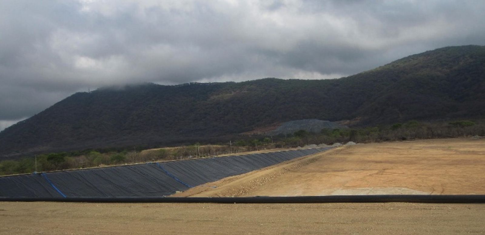 A geomembrane-lined embankment slope running along the edge of a prepared basin under a cloudy sky, with a mountain behind