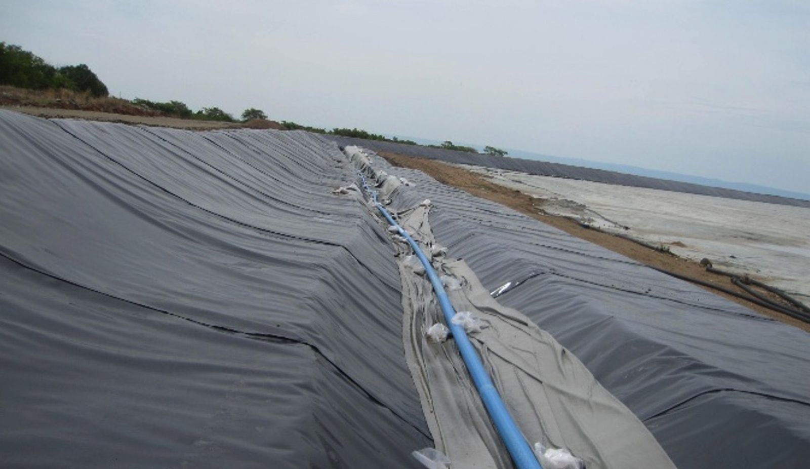 Rolled and overlapped geomembrane sheets lining a long embankment slope, with a drainage pipe running along the seam