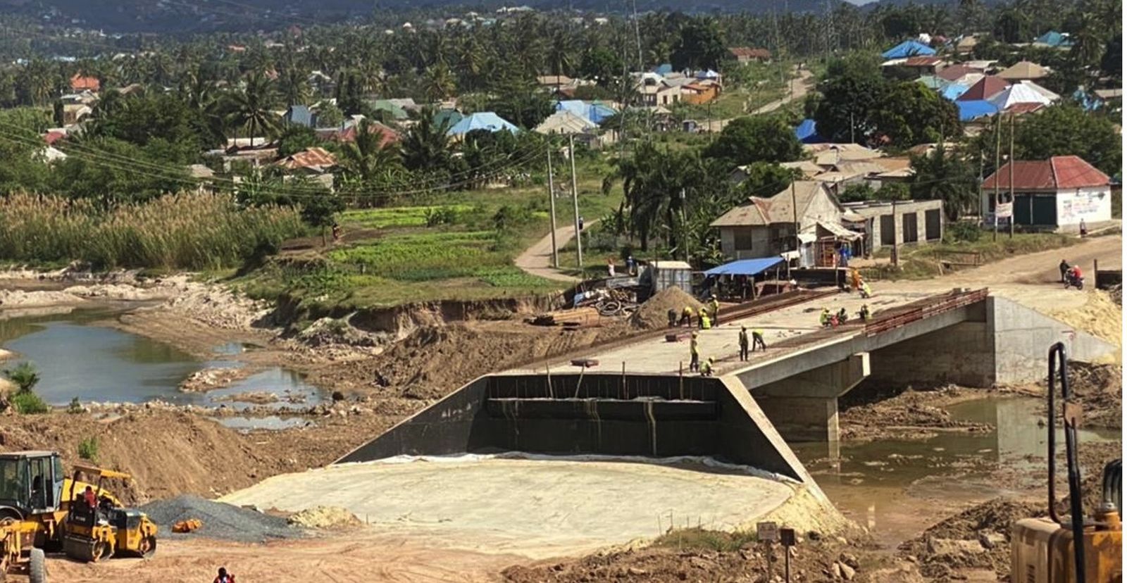 A reinforced concrete bridge under construction across a sandy river channel, with a concrete apron, workers on the deck, a roller and a loader, and a densely built hillside settlement behind