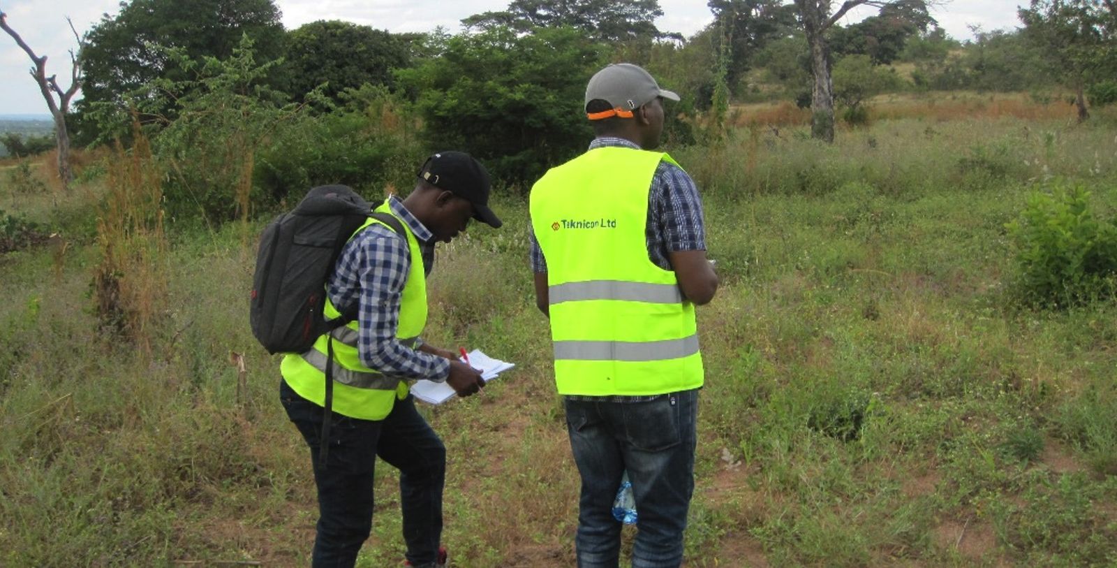 Two engineers in yellow high-visibility vests, one printed Teknicon Ltd, taking notes on a clipboard during a route survey in open bush country