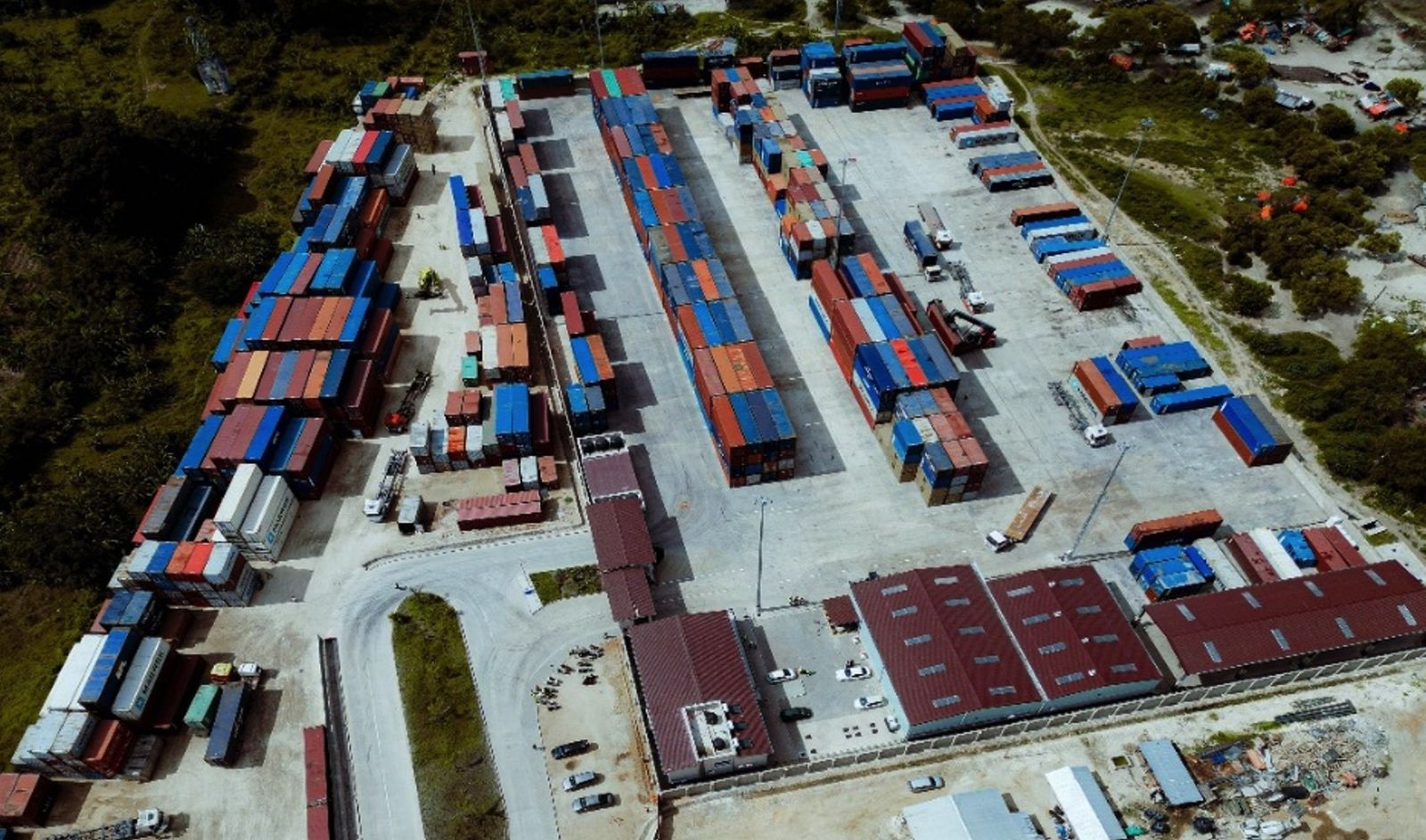 Drone view of a container storage yard with rows of stacked shipping containers on a paved hardstand, alongside red-roofed workshop and service buildings